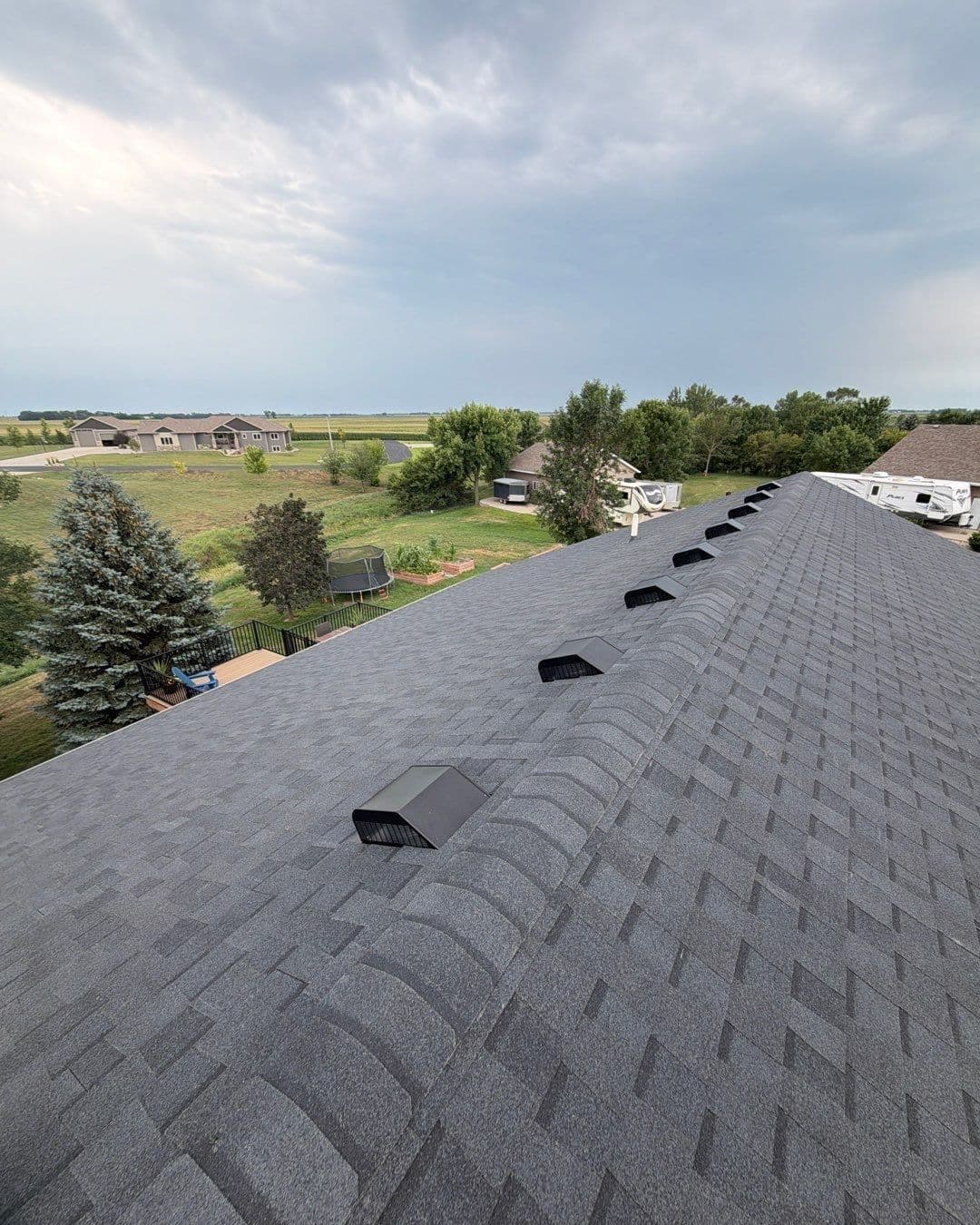 Aerial view of a roof with multiple black attic vents, overlooking a rural property with farm buildings, trees, and fields under a cloudy sky
